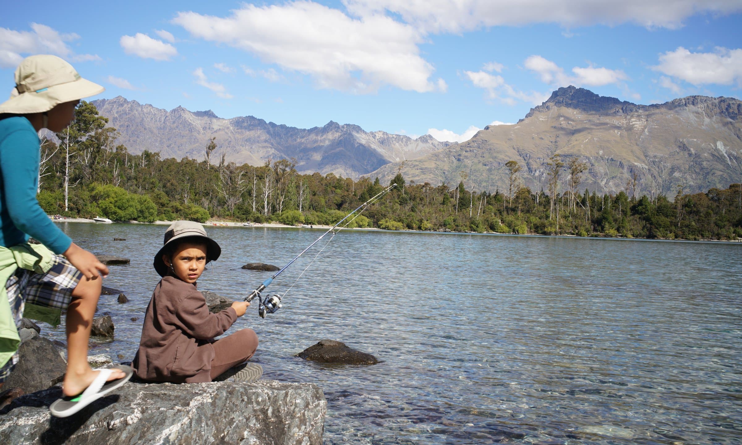 Reyaan fishing on the rocks by a lake, with mountains in the background